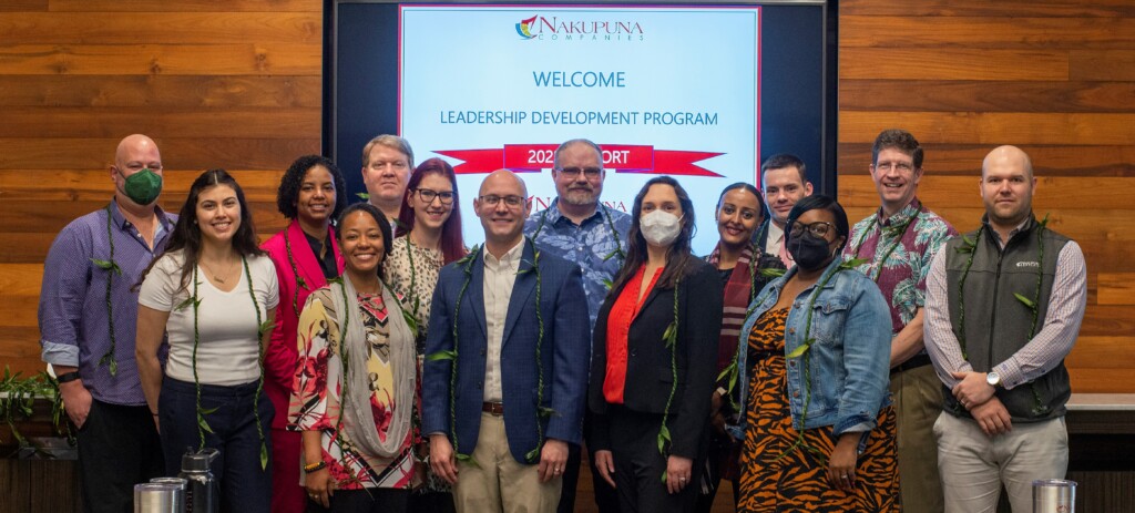 Nakupuna Companies' 2023 Leadership Development Program graduates pose for a group photo. The diverse group of smiling professionals wear leis and stand in front of a welcome banner. This image showcases career development opportunities at Nakupuna Companies.
