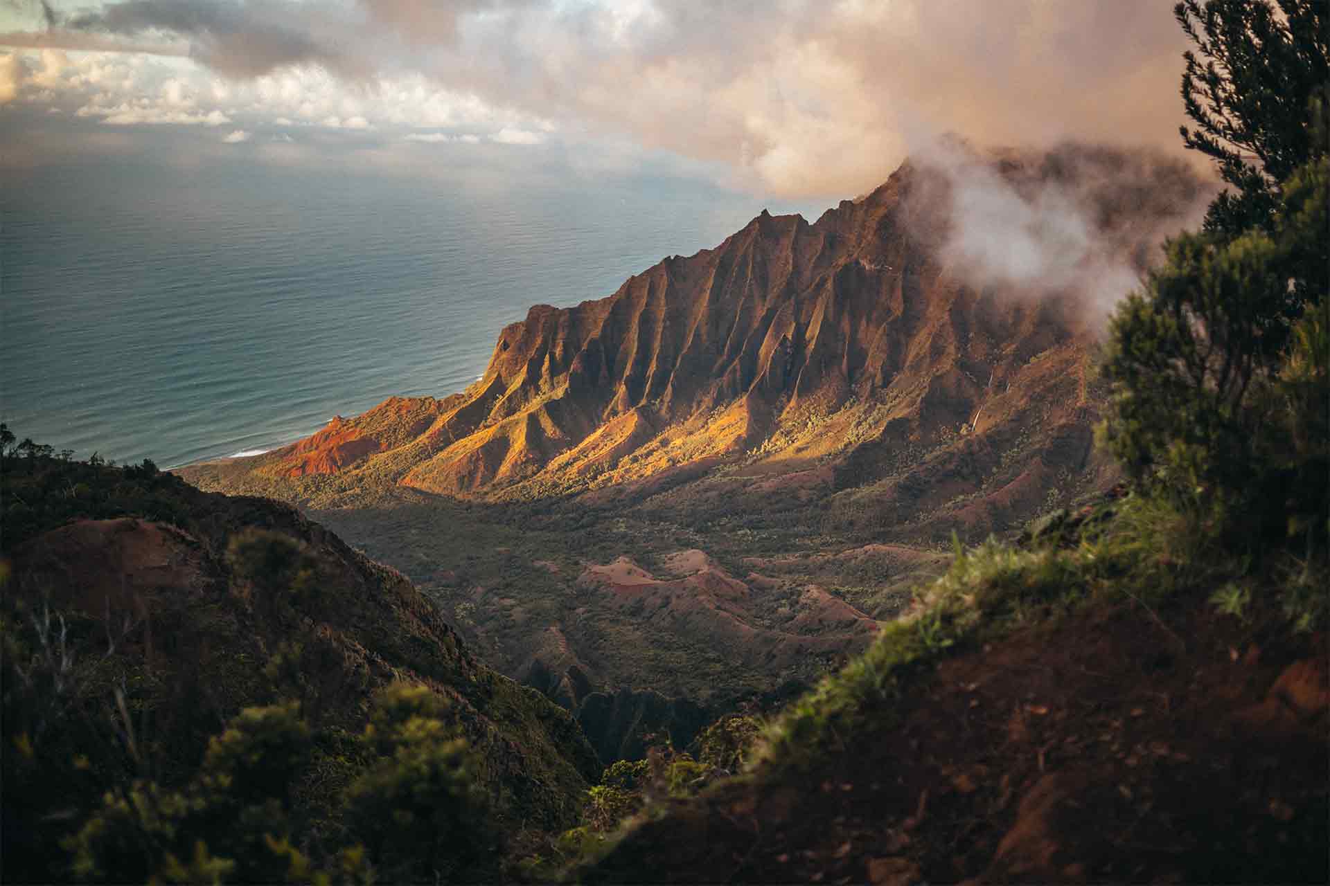 Dramatic Na Pali Coast cliffs on Kauai bathed in golden sunset light, with clouds rolling in from the Pacific Ocean and lush green valleys below. This image is featured on the leadership pages of the Nakupuna Companies website.