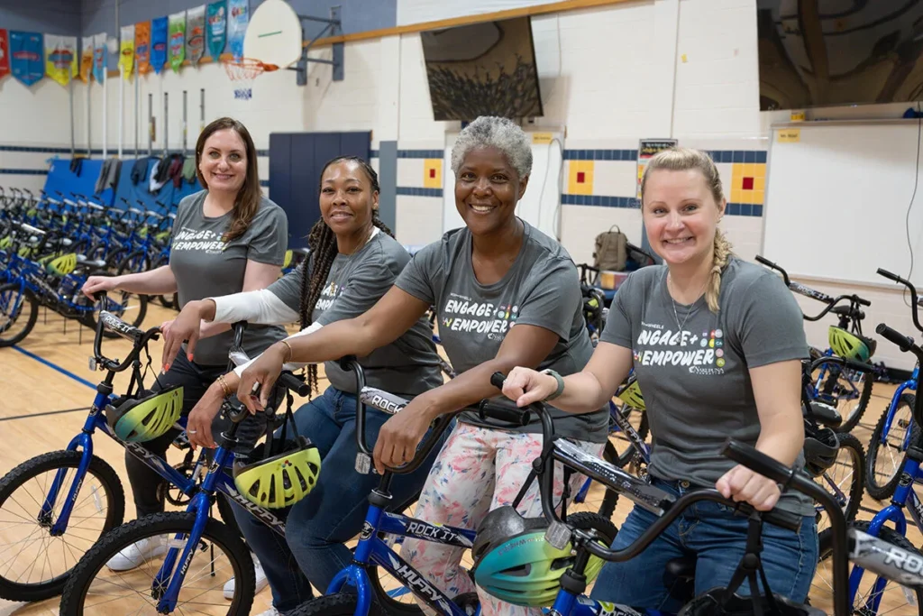 2025 Wish For Wheels Arlington - Project Kupu Four smiling women pose with blue Huffy bikes and helmets in a gym, wearing "Engage Empower" shirts. They are volunteering for the Nakupuna Companies Project Kupu Wish for Wheels event in Arlington.