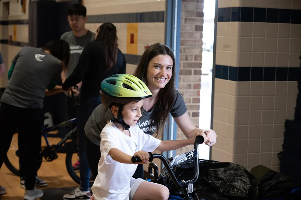 2025 Wish For Wheels Arlington - Project Kupu A smiling woman helps a young student with a yellow helmet ride a bike indoors. This image was taken at the Project Kupu Wish for Wheels and Nakupuna Foundation collaborative event at the Arlington location for Nakupuna Companies.