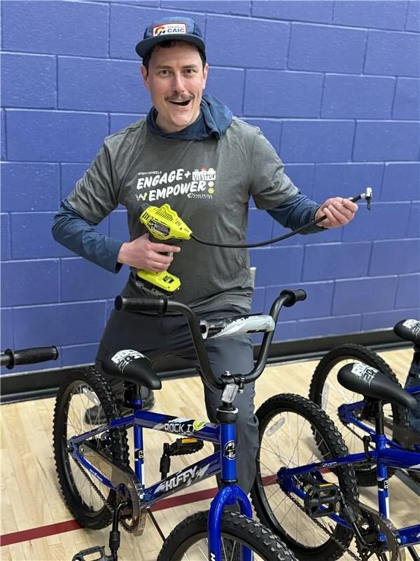 2025 Wish For Wheels Arlington - Project Kupu Man with a mustache pumps a Huffy BMX bike tire with a Ryobi power tool. He wears a grey "Engage + Empower" Wish for Wheels t-shirt and a CAIC hat in a gymnasium. Bikes are lined up behind him. This image was taken at the Project Kupu Wish for Wheels and Nakupuna Foundation collaborative event at the Arlington location for Nakupuna Companies.