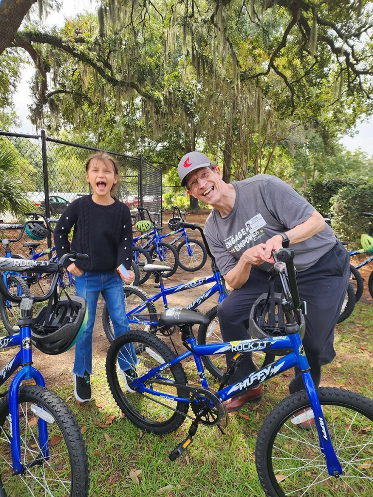 2025 Wish For Wheels Charleston - Project Kupu A happy student and man pose with new Huffy bikes outdoors. The man wears an "Engage, Empower" Wish for Wheels shirt and a Nakupuna Companies cap. Blue bikes stand ready for kids in a park setting with hanging moss. This image was taken at the Project Kupu Wish for Wheels and Nakupuna Foundation collaborative event at the Charleston location for Nakupuna Companies.