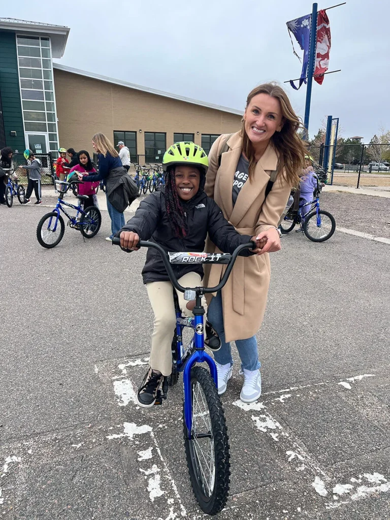 2025 Wish For Wheels Denver - Project Kupu A young student with a bright smile learns to ride a blue bicycle at the Wish for Wheels Project Kupu event in Denver. A Nakupuna employee in a tan coat holds the bike steady, smiling warmly. Other students, employees and bikes are visible in the schoolyard background.