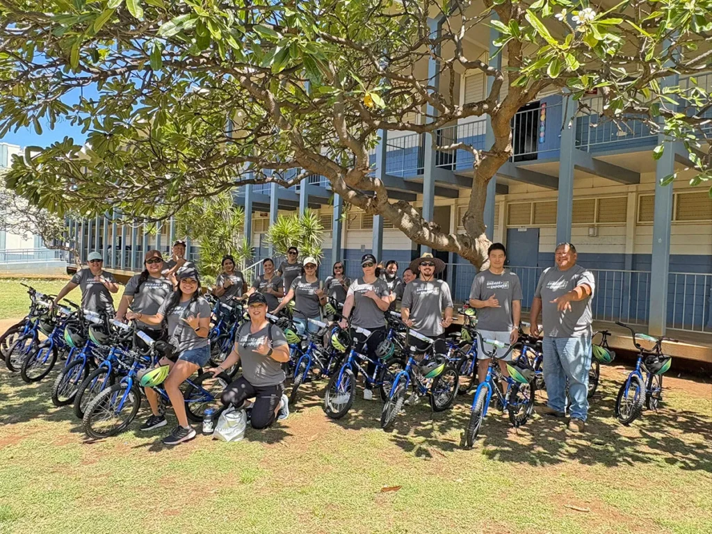 2025 Wish For Wheels Honolulu - Project Kupu Nakupuna employees pose with newly assembled blue bicycles under a large tree in front of a blue and white building. This image was taken at the Project Kupu Wish for Wheels and Nakupuna Foundation collaborative event at the Hawai‘i location for Nakupuna Companies.