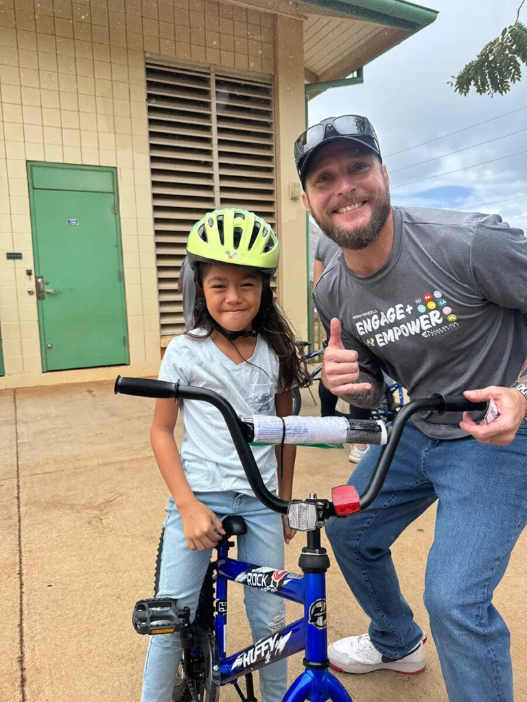 2025 Wish For Wheels Honolulu - Project Kupu A smiling Nakupuna employee gives a thumbs up next to a young girl on a blue Huffy bicycle with a yellow helmet. The man wears a gray "Engage + Empower" t-shirt with the Wish for Wheels and Nakupuna Foundation logos. This image is featured on the Nakupuna Companies website.