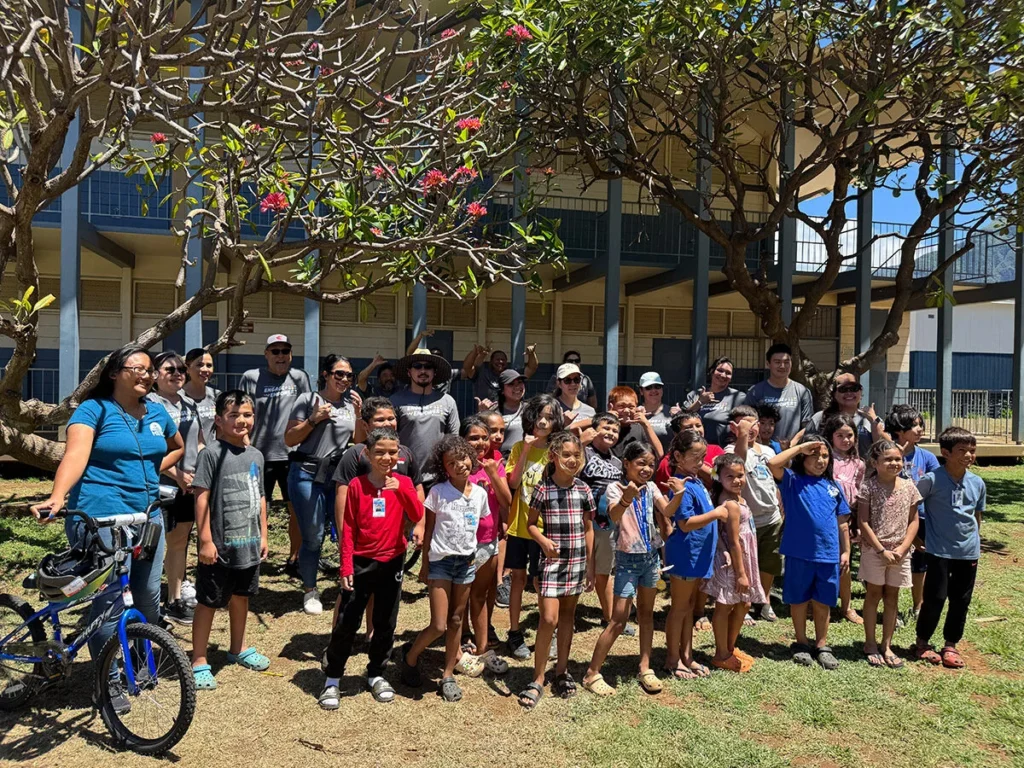2025 Wish For Wheels Honolulu - Project Kupu A group of students and Nakupuna employees stand together smiling in front of a school building, framed by flowering trees. A blue bicycle leans against a person on the left. The mood is sunny and cheerful. This image was taken at the Project Kupu Wish for Wheels and Nakupuna Foundation collaborative event at the Hawai‘i location for Nakupuna Companies.