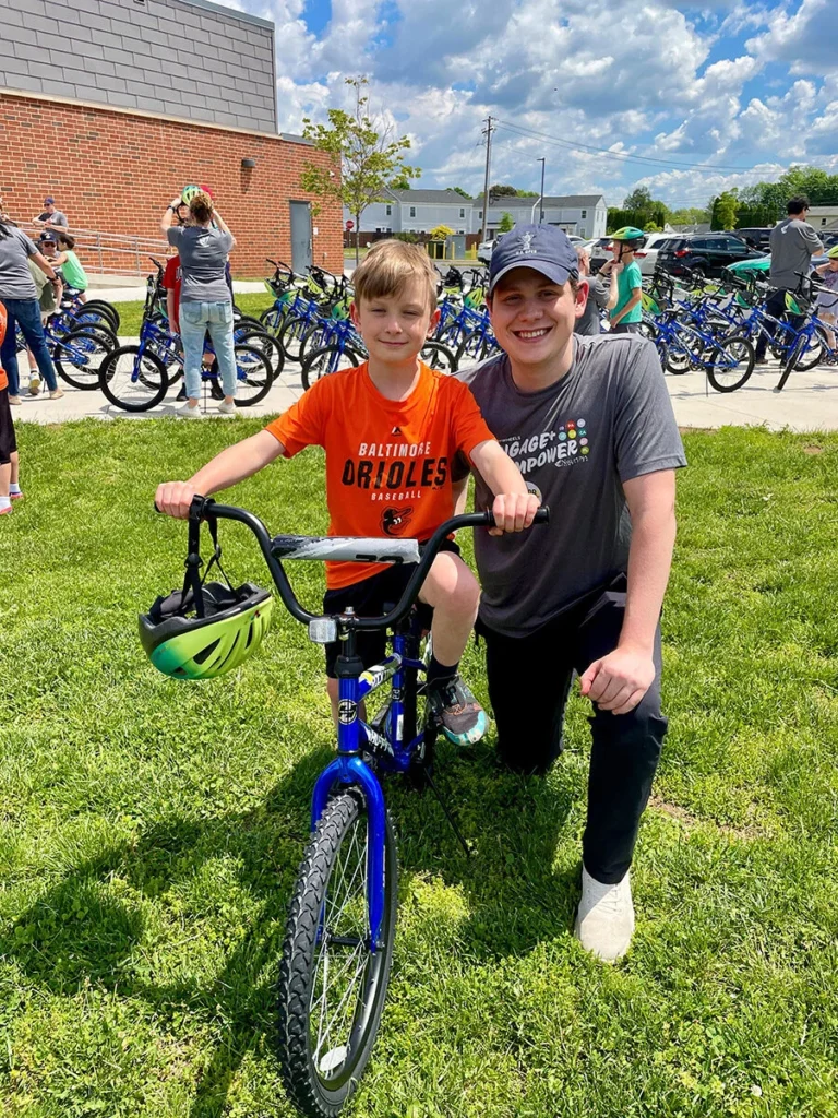 2025 Wish For Wheels Mechanicsburg - Project Kupu Student in an orange Baltimore Orioles shirt smiles on a new blue bike provided by Wish for Wheels and Nakupuna Foundation as part of their Project Kupu charity event in Mechanicsburg. Standing next to the student is a Nakupuna Companies employee.
