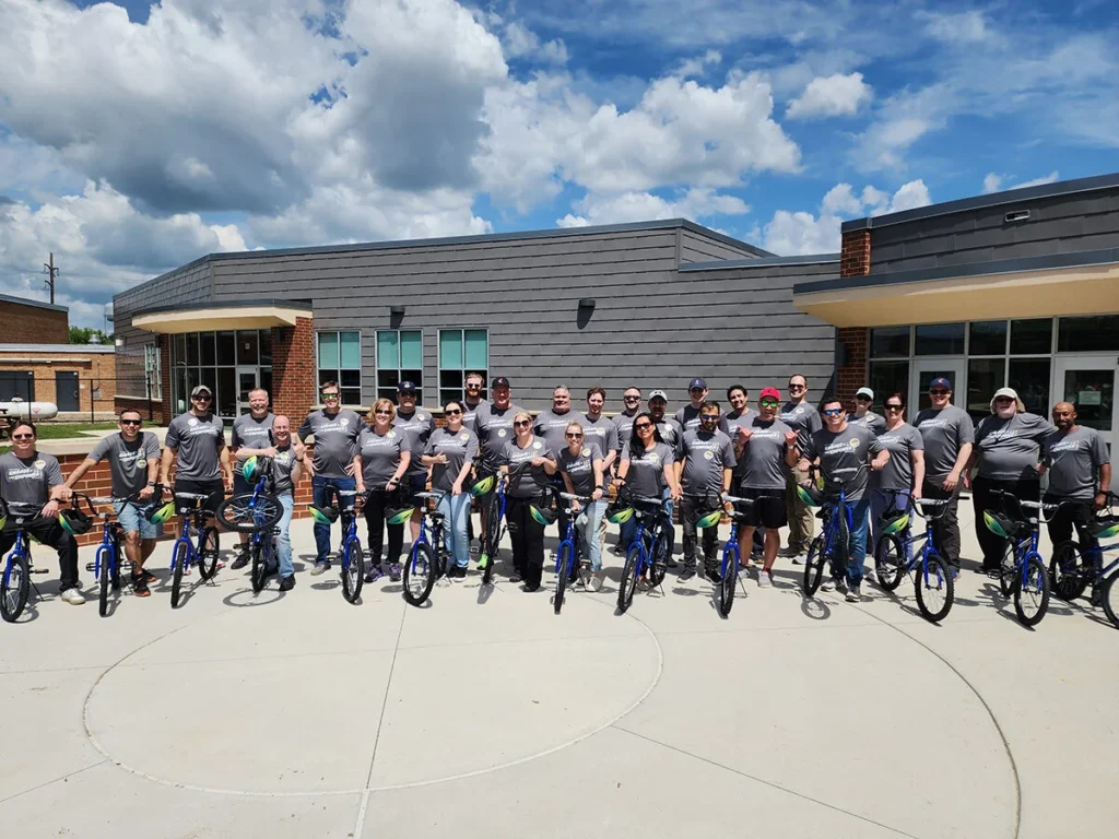 2025 Wish For Wheels Mechanicsburg - Project Kupu Nakupuna Employees at the Mechanicsburg office stand with blue bicycles in front of a modern building under a partly cloudy sky for the Project Kupu Wish for Wheels event.. They wear matching gray shirts with "Engage + Empower" written on them along with the Wish for Wheels and Nakupuna Foundation logos.