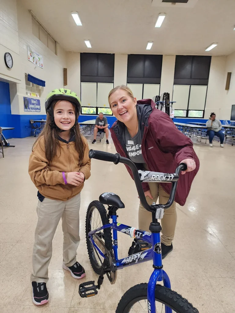 2025 Wish For Wheels Norfolk - Project Kupu A smiling young girl in a helmet stands with a Nakupuna Companies employee next to a new blue Huffy bicycle in a brightly lit gymnasium at a Project Kupu Wish for Wheels event in Norfolk.