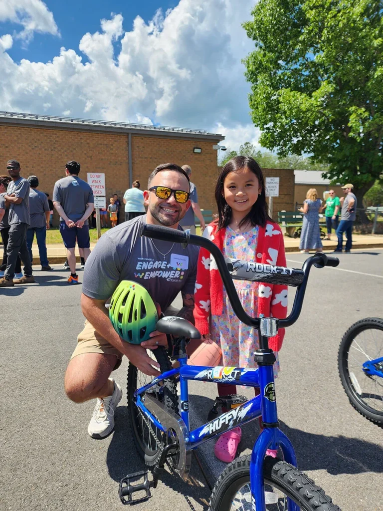 2025 Wish For Wheels Pax River - Project Kupu A smiling student stands with Nakupuna Companies employee, kneeling next to a blue Huffy bicycle and a green helmet. The Wish for Wheels Pax River event is outdoors on a sunny day with other people in the background.