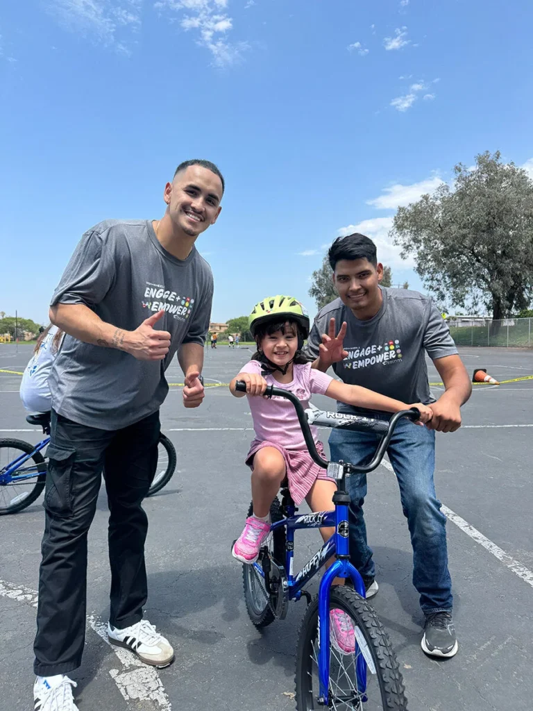 2025 Wish For Wheels San Diego - Project Kupu A young student beams on a blue bicycle, wearing a yellow helmet, flanked by two smiling Nakupuna Companies volunteers at the San Diego Wish for Wheels event. They are giving thumbs up and peace signs in an outdoor parking lot setting.