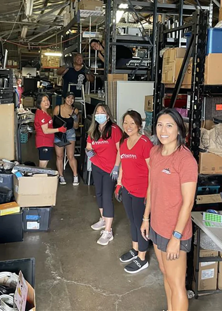 Hawaiian Hope Project Kupu Nakupuna Companies 2 Volunteers in red shirts and face masks sort through e-waste in a warehouse. Shelves full of electronics and boxes line the background. The shirts say "DYWAM". This image is featured on the Nakupuna Companies website.