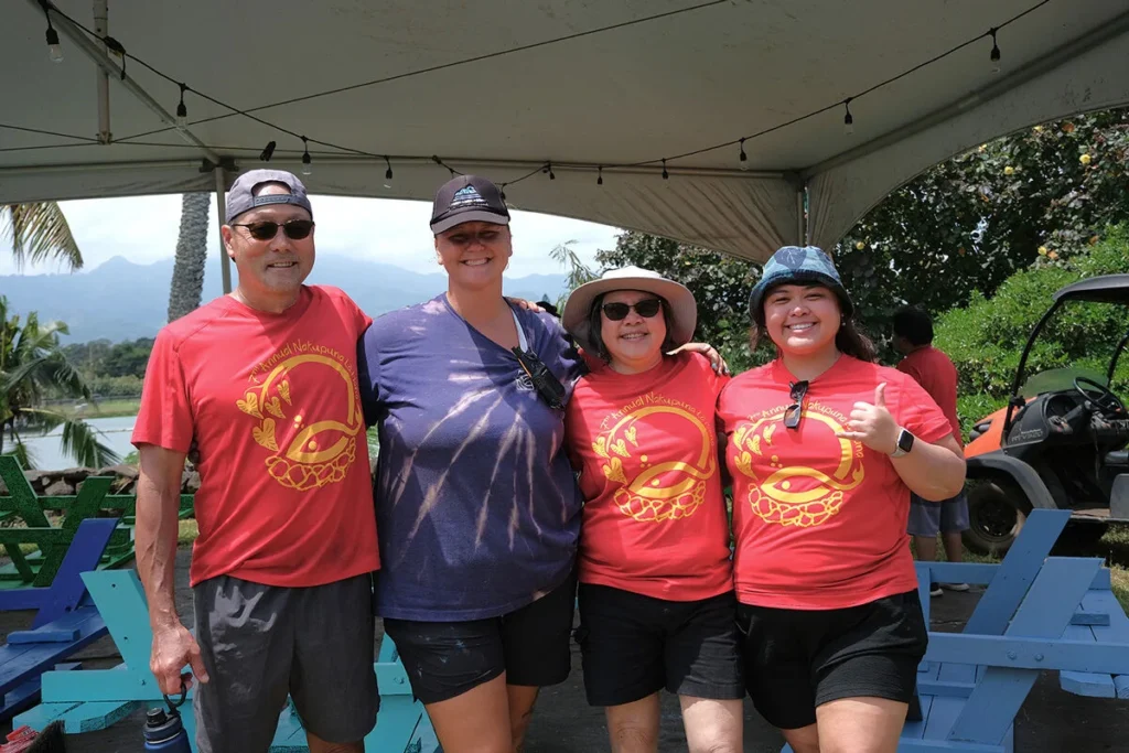 Nakupuna employees pose under a tent with string lights. Three wear red shirts with a gold design; one gives a thumbs up. Lush greenery, mountains, and a golf cart are visible in the background. This image is featured on the Nakupuna Companies website.
