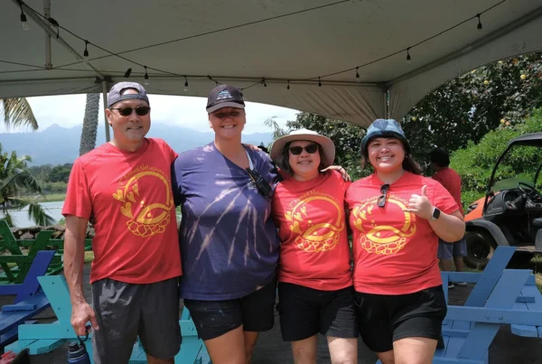 Nakupuna employees pose under a tent with string lights. Three wear red shirts with a gold design; one gives a thumbs up. Lush greenery, mountains, and a golf cart are visible in the background. This image is featured on the Nakupuna Companies website.