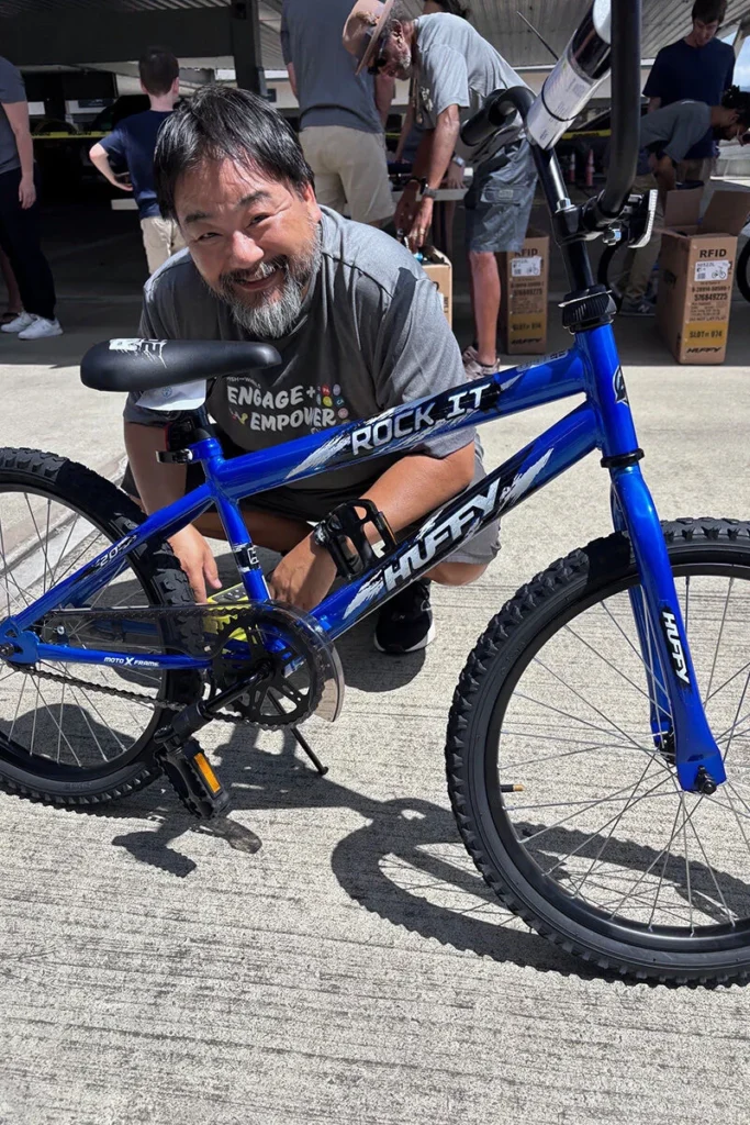 Wish For Wheels Project Kupu Nakupuna Companies 2 A smiling Asian man kneels next to a new blue Huffy Rock It BMX bike. The bike features black tires, a kickstand, and "Engage + Empower + Rock It" text on the frame. Boxes labeled "RFID" are in the background. This image is featured on the Nakupuna Companies website.