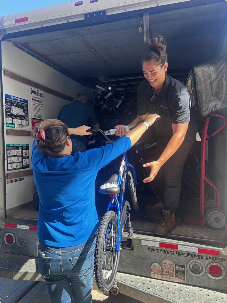 Wish For Wheels Project Kupu Nakupuna Companies 3 Two people unload a truck filled with bicycles. One person in a blue shirt hands a blue bicycle to another person in a black shirt, who is smiling. The truck is packed with bikes, ready for donation. This image is featured on the Nakupuna Companies website.