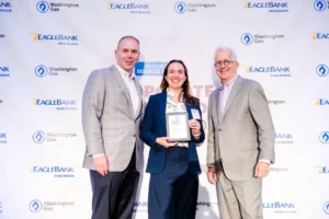 Three professionals pose for a photo at the Washington Business Awards. A woman in a navy blazer holds an award. The background features the event's sponsors, Washington Gas and EagleBank. This image is featured on the Nakupuna Companies website.
