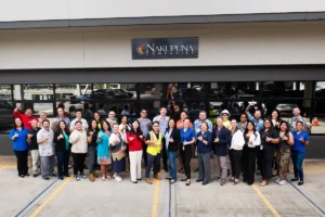Nakupuna Companies team celebrates WTOP Top Workplace award outside their building. Diverse employees in casual attire smile broadly in front of the Nakupuna Companies logo, a government contractor recognized for its employee engagement. 