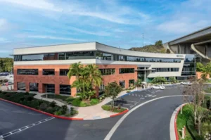 Modern office building with a curved facade and red brick accents. Lush palm trees and landscaping surround the building under a partly cloudy sky, with a highway overpass in the background.