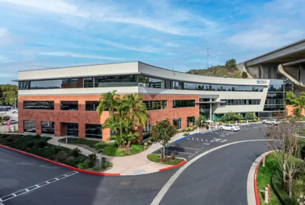 Modern office building with a curved facade and red brick accents. Lush palm trees and landscaping surround the building under a partly cloudy sky, with a highway overpass in the background.