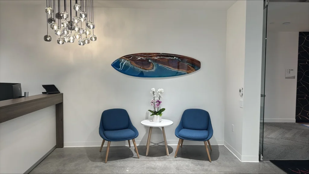 Nakupuna Companies - San Diego Entrance Seating A modern waiting area features two blue upholstered chairs facing a small white table with a potted orchid, beneath a striking oval surfboard shaped resin art piece resembling a wave. A contemporary chandelier hangs above.