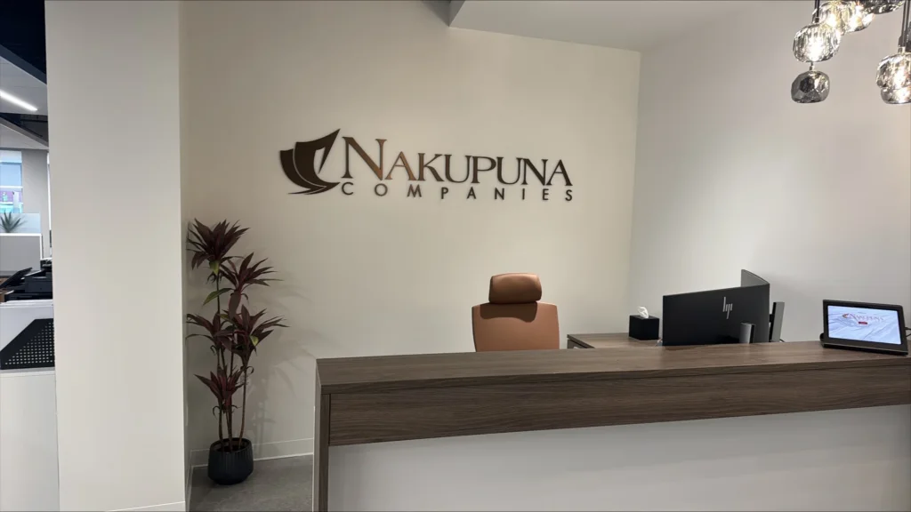 Nakupuna Companies - San Diego Entrance Nakupuna Companies logo prominently displayed on a modern office reception wall above a sleek wooden desk with a computer and tablet. A potted plant adds a touch of greenery to the professional space.